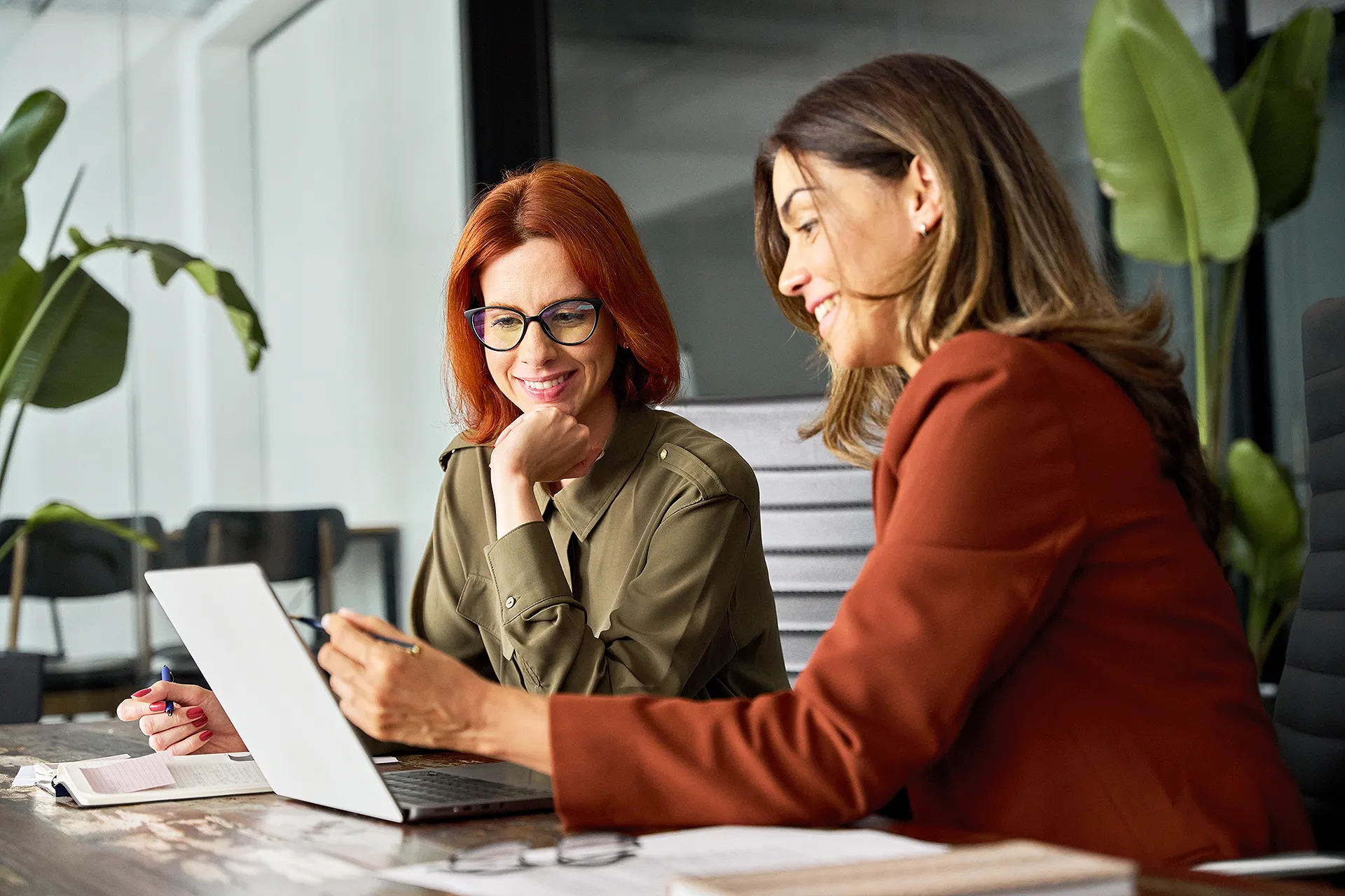 Middle aged professional business woman consulting teaching young employee looking at laptop sitting at desk in office